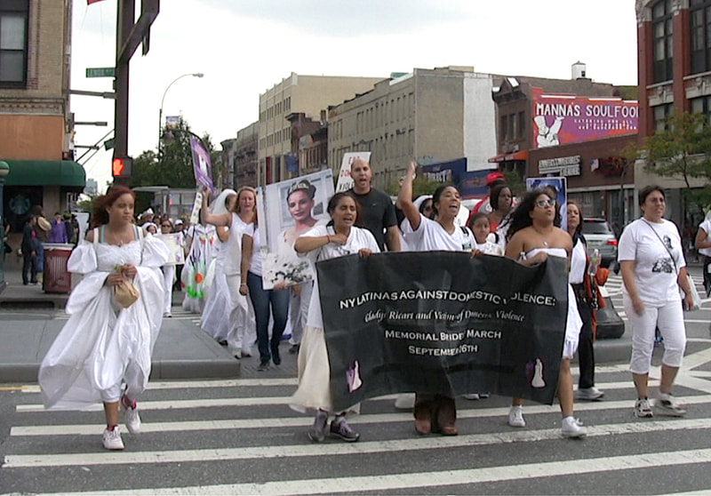 Women dressed in wedding gowns marching behind a black banner.