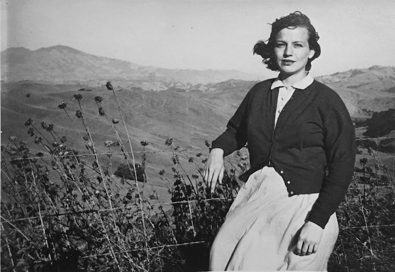 Black and white photo of a young white woman standing in front of a vista of hills and mountains.
