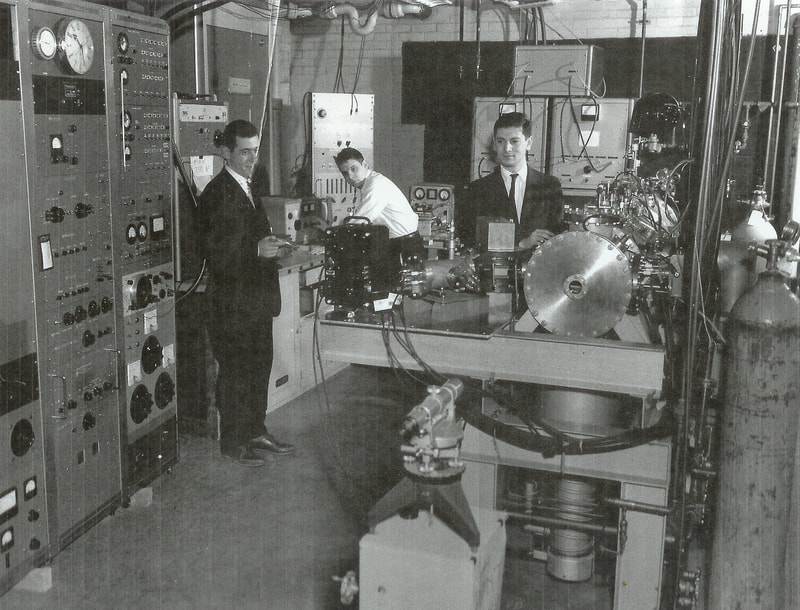 Black and white photo of three white men in a laboratory with large machines. 