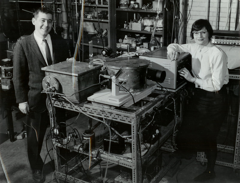 Black and white photo of a young white man and a young white woman standing beside laboratory equipment.