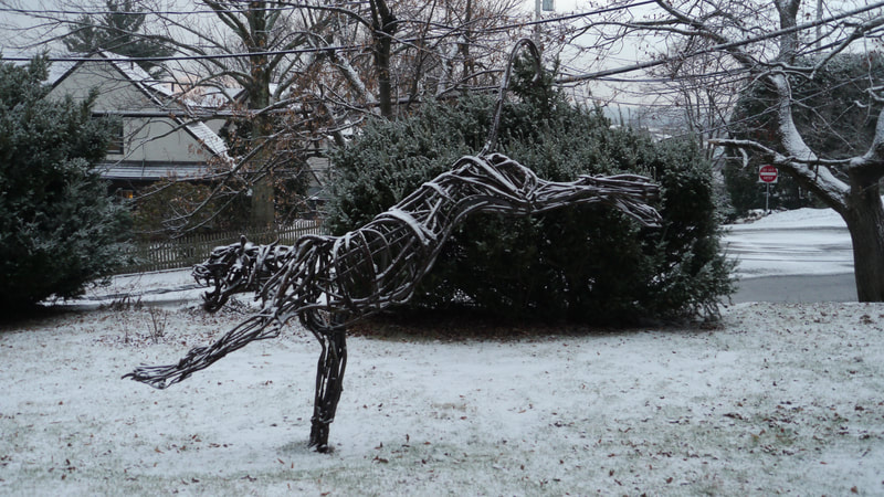 A large welded steel sculpture of a leaping tiger on a snow-covered lawn. There are trees and utility wires in the background.