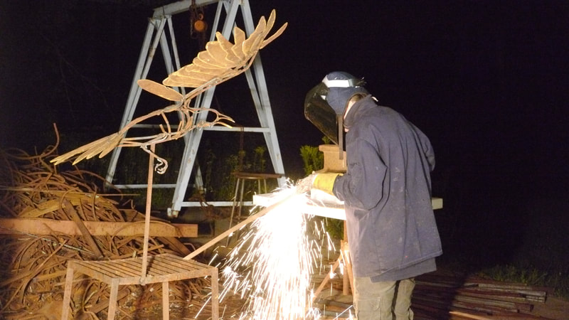 A Person in a helmet and long jacket cutting metal at night next to a metal bird sculpture.